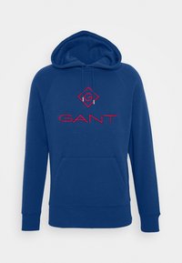 La sudadera azul presenta un bolsillo frontal, capucha con cordones y un logo bordado en rojo con el texto "GANT", confeccionada en una tela suave.