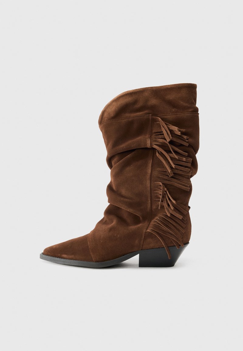 Bota de tornozelo em camurça castanha com um design descaído, bico fino e detalhes de franjas ao longo do lado exterior. Salto empilhado para um maior comprimento.