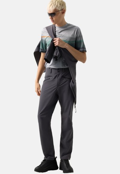 Junge Person mit Sonnenbrille, grafischem T-Shirt, dunkler Hose und schwarzen Schuhen, hält eine dunkle Jacke über eine Schulter gelegt.