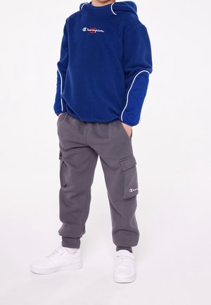 Kind trägt einen blauen Champion-Hoodie und graue Cargo-Sweathosen mit weißen Turnschuhen, steht mit den Händen in den Taschen vor weißem Hintergrund.