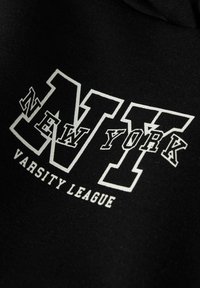 Sweat à capuche noir avec un grand texte blanc indiquant "NEW YORK" au-dessus de "VARSITY LEAGUE", dans un design de police universitaire audacieuse.