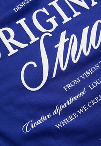 Μπλε βαμβακερό T-shirt με μεγάλα λευκά γράμματα σε συνδυασμό έντονων και καλλιγραφικών γραμματοσειρών. Ο τίτλος περιλαμβάνει τις λέξεις "Πρωτότυπο" και "Δημιουργικό τμήμα."