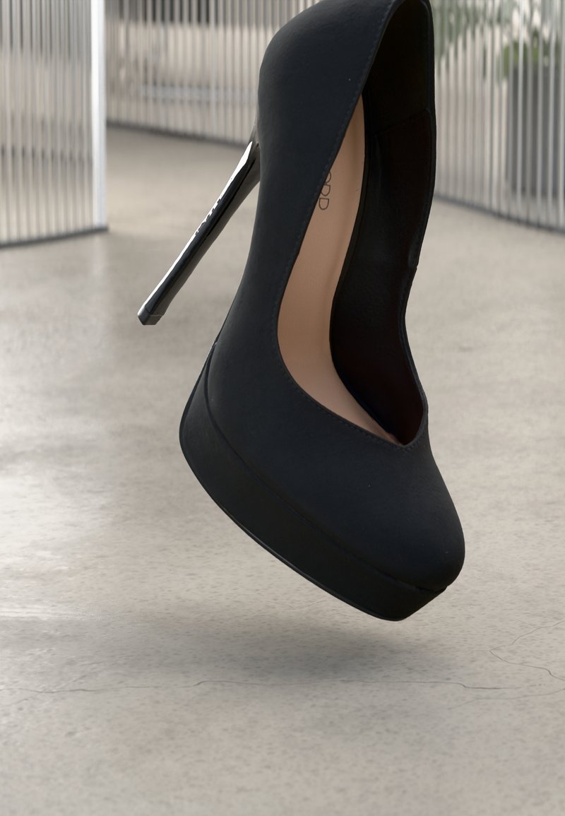 Chaussure à plateforme noire à talons aiguilles avec un bout pointu, présentée sur un sol gris texturé et un arrière-plan de barres verticales métalliques.