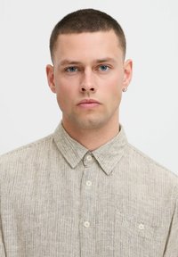 Beige stribet skjorte med knaplukning, spidse krave, en enkelt brystlomme og cremefarvede knapper. Stoffet har en tekstureret overflade.