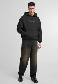 Crna dukserica s kapuljačom koja ima prednji džep i bijeli natpis. Uparena s širokim crnim trapericama i crnim cipelama.