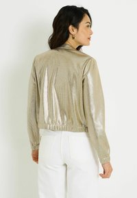 Blouson aviateur doré métallisé avec taille et poignets élastiques, présentant une texture lisse et une finition brillante. Porté avec un pantalon blanc.