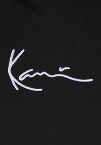 Čierna látka so šitým podpisovým logom v bielej farbe, ktoré má hladké krivky a rôznu hrúbku čiar.