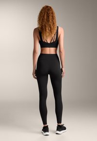 Črn športni modrček z več zadnjimi trakovi, v kombinaciji s črnimi visokimi leginami. Material je raztegljiv in zasnovan za športno uporabo.