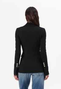 Zwarte getailleerde blazer met een gladde textuur, een slim fit ontwerp en gouden knoopdetails op de mouwen, gedragen over lichtblauwe jeans.