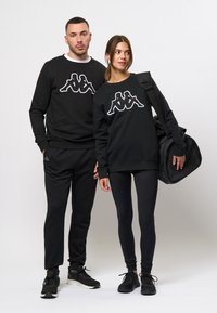 Zwarte sweatshirts hebben een wit logo op de borst. De man draagt een bijpassende zwarte broek; de vrouw draagt zwarte leggings en neemt een zwarte sporttas mee.