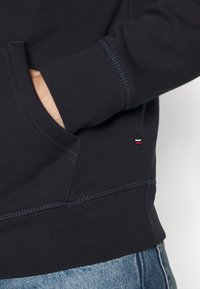 Marineblaues Sweatshirt mit gerippter Textur, ausgestattet mit einer Kängurutasche, kontrastierenden Nähten und einem kleinen Logosticker in Rot, Weiß und Schwarz.