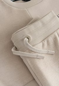 Bežs kokvilnas maisījuma džemperis un saskaņoti sporta bikses, ar strukturētu audumu, ribotām aprocēm un auklu ar metāla akcentiem.