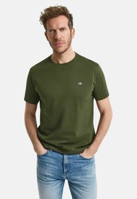 Tricou din bumbac de culoare verde măsliniu, cu guler rotund și mâneci scurte, având un logo mic în partea stângă a pieptului. Purtați cu blugi albaștri.