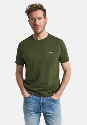 Olive green cotton T-shirt with a crew neckline and short sleeves, featuring a small logo on the left chest area. Worn with blue jeans.