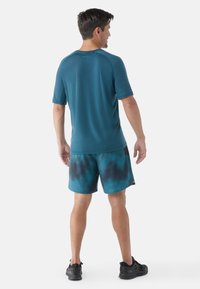 Teal sportovní tričko s krátkými rukávy zkombinované s řasenými šortkami modrého tie-dye. Černé akcenty na šortkách. Model má na sobě černé sportovní boty.