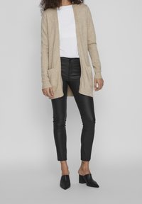 Beige lång cardigan med ribbad mudd, framfickor och öppen design framtill, parat med svarta tajta byxor och svarta klackskor.