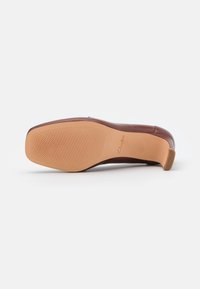 Scarpa con tacco alto in pelle marrone, dalla superficie liscia, con una suola in gomma beige chiaro caratterizzata da un motivo di grip testurizzato. Accento su un tacco piccolo.