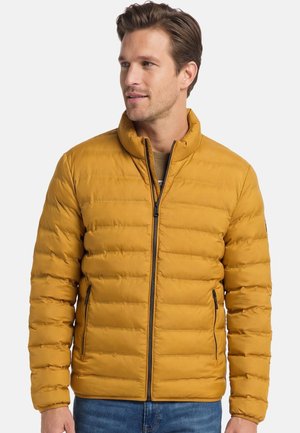 Mand iført sennepsgul quiltet jakke med lynlås foran og lynlåslommer, der kigger let mod højre mod ensartet baggrund.