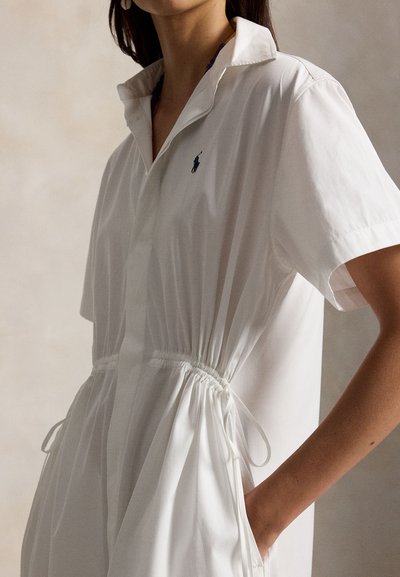 Femme portant une robe blanche à manches courtes avec un col, une taille ajustable cintrée et un petit logo brodé sur la poitrine.