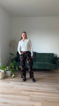 Bela srajca s ovratnikom, črne široke hlače s cvetličnim vzorcem in črne čevlje. Preprosto pohištvo in rože v svetli sobi.