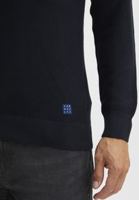 Mornarska rebrasta pletena jopica s zaobljenim robom, ki ima na levi strani modro oznako z belim napisom "DNWORKRS". Dolgi rokavi in oprijete manšete.