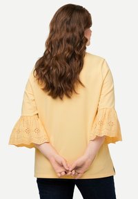 Top di cotone giallo con maniche a sbuffo in pizzo a occhio, corpo solido, spacchi laterali e orlo arrotondato, abbinato a jeans blu scuro.