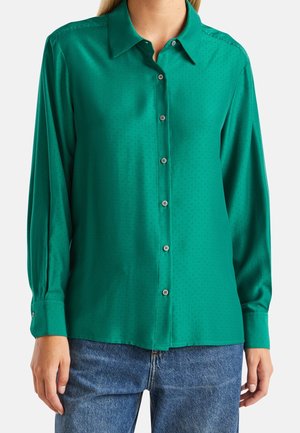Chemise en satin vert à manches longues, col classique, motif à pois subtil et poignets à boutons. Associée à un jean en denim bleu.