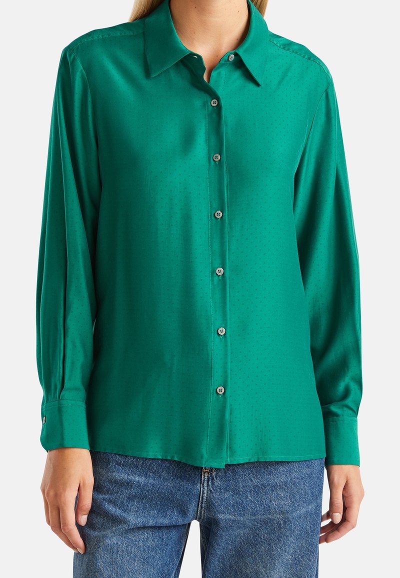 Grön satinknappad skjorta med långa ärmar, klassisk krage, subtila prickar och knäppta manschetter. Tillsammans med blå denimjeans.