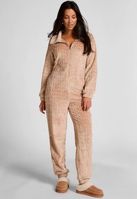 Beige myk koseoverall med glidelås, med myk tekstur, høy krage, lange ermer og quiltet mønster; kombinert med komfortable slip-on sko.