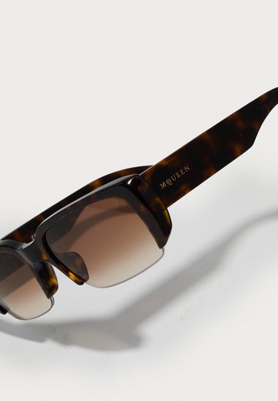 Occhiali da sole tartarugati con lenti marroni sfumate, montatura quadrata oversize e logo dorato "McQueen" sulla tempia.