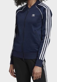 Osoba v námořnické modré bundě Adidas s bílými pruhy na rukávech a se zapínacími kapsami, doplněná o černé kalhoty s bílými pruhy.