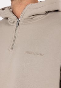 Sweat à capuche beige avec fermeture éclair, finition texturée, doté d'une fermeture métallique et d'un logo discrètement embossé sur la poitrine. Tissu lisse avec capuche.