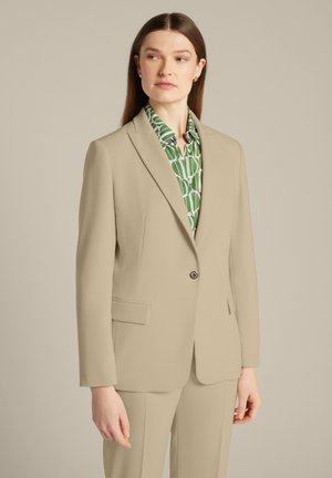 Beiger Blazer mit strukturierten Schultern, einem einzigen Knopfverschluss und vorderen Taschen, kombiniert mit einem grünen gemusterten Hemd darunter.