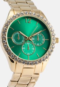 Montre-bracelet dorée avec un cadran rond vert et trois sous-cadrans, entourée d'une lunette ornée de cristaux et d'un bracelet en métal doré.