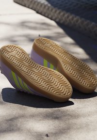 Par lilla og neon-gule sneakers liggende med sålerne opad på en solbeskinnet betonoverflade med synlige teksturerede gummiydersåler.