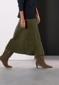 Knit, olive green skirt with textured ribbing, paired with brown, knee-high, pointed boots featuring side zippers. Model walking.