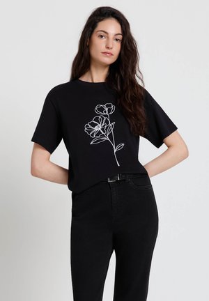 Femme aux longs cheveux foncés portant un t-shirt noir avec un dessin floral blanc en lignes et un jean noir taille haute, debout devant un fond clair.