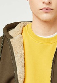 Brun hoodie med en beige, blød, pelsforet inderside, lagd oven på en ensfarvet gul sweatshirt med ribbet krave.