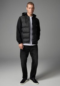 Gilet rembourré noir sur un t-shirt à manches longues noir, associé à un jean foncé et des baskets noires. Gilet zippé avec capuche et texture lisse.