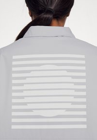 Chaqueta gris con un gran diseño gráfico en la parte posterior, que consiste en líneas horizontales y una forma circular en blanco. Acabado textil suave.