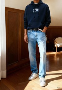 Homme portant un sweat à capuche bleu marine avec un logo, un jean bleu clair, une ceinture noire et des baskets blanches, debout sur un sol en bois dans une pièce ensoleillée.