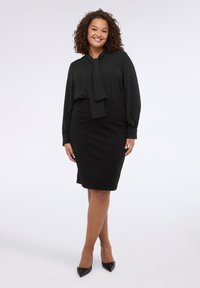 Bluza neagră cu detaliu de guler legat și mâneci lungi, asociată cu o fustă creion neagră, mulată și pantofi negri cu vârf ascuțit. Textură strălucitoare.