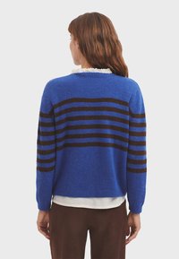 Blauer Pullover mit braunen horizontalen Streifen, aus einem weichen Material, mit Rundhalsausschnitt und lockerer Passform. Akzentuiert mit einem weißen Hemd darunter.