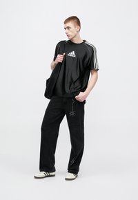 Μαύρο αθλητικό μπλουζάκι με λευκές ρίγες και λογότυπο, συνδυασμένο με μαύρο τζιν με φθορές και λεπτομέρειες με τρουκς. Τα παπούτσια είναι λευκά με ρίγες.