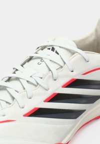 Λευκό αθλητικό παπούτσι με γκρι και μαύρες αποχρώσεις, με τρεις μαύρες ρίγες και κόκκινες λεπτομέρειες. Λείο δερμάτινο επάνω μέρος και υφασμένο πέλμα.