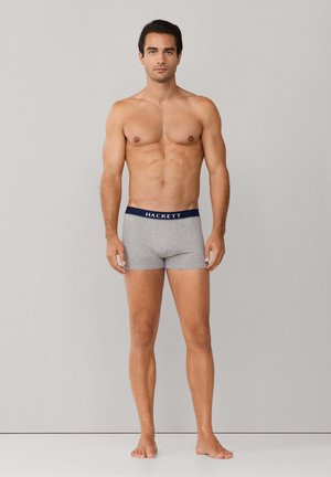 Un homme musclé se tient pieds nus, portant un boxer gris avec une ceinture bleu marine marquée "HACKETT", sur un fond clair uni.