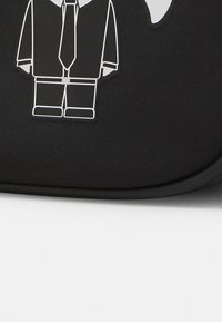 Sac noir avec un graphique d'un personnage de dessin animé en costume noir avec une cravate, tracé en blanc, sur un fond texturé.