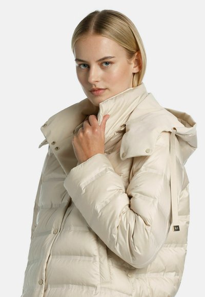 Beige pufferjack met een hoge kraag, drukknoopsluiting en trekkoorddetails. Het materiaal is glad en heeft een gewatteerde textuur voor extra warmte.