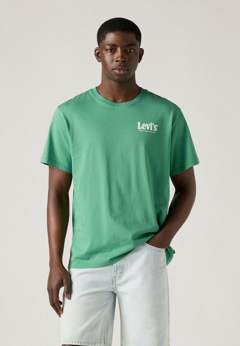 Junger Mann, der ein grünes Levi's T-Shirt und helle Denim-Shorts trägt, steht mit einer Hand in der Tasche vor einfachem Hintergrund.