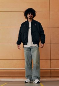 Musta bomber-takki valkoisen t-paidan päällä, kulutetut vaaleansiniset farkut ja mustavalkoiset tennarit; teksturoitu parta ja aurinkolasit.